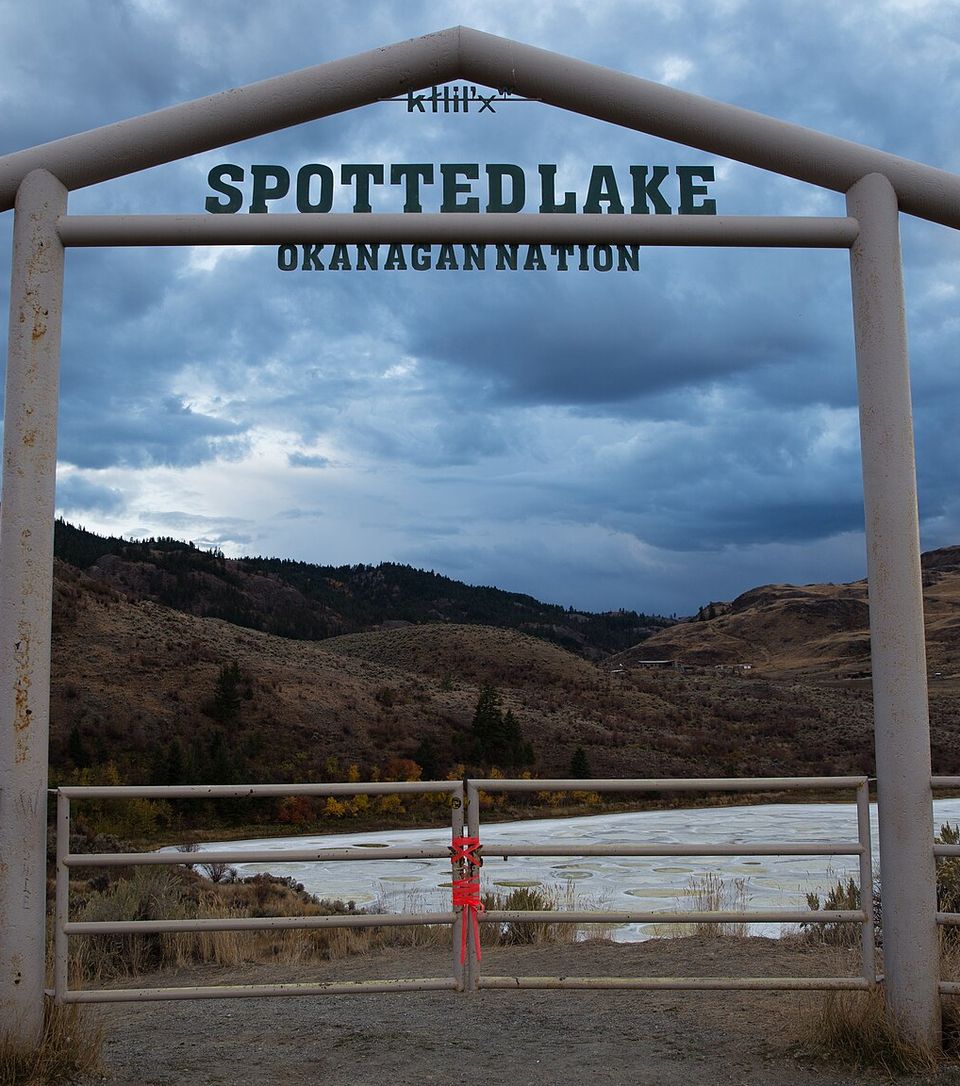 A white pipe arch reading "Spotted Lake - Okanagan Nation" over a padlocked white gate tied with a red ribbon, the spotted lake pale beyond under an evening sky