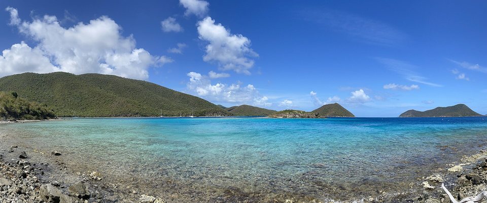 A wide clear turquoise bay with a rocky shore in front and forested green hills and a small cay across the water