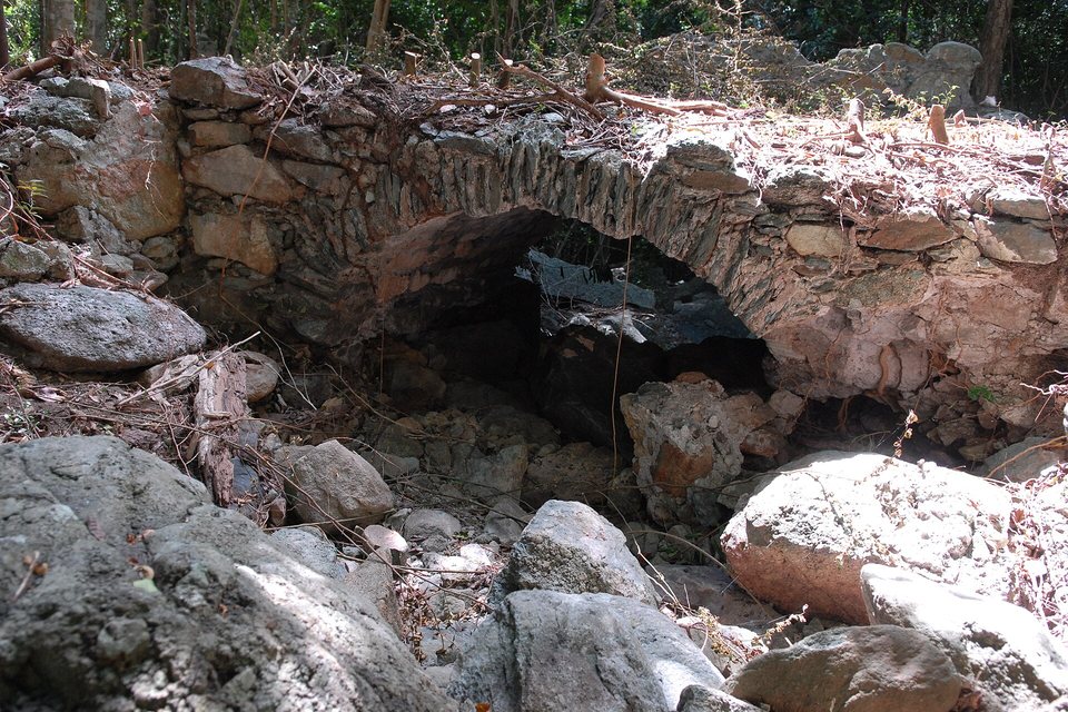 A low stone arch bridge over a dry rocky streambed, covered in leaf litter and fallen branches