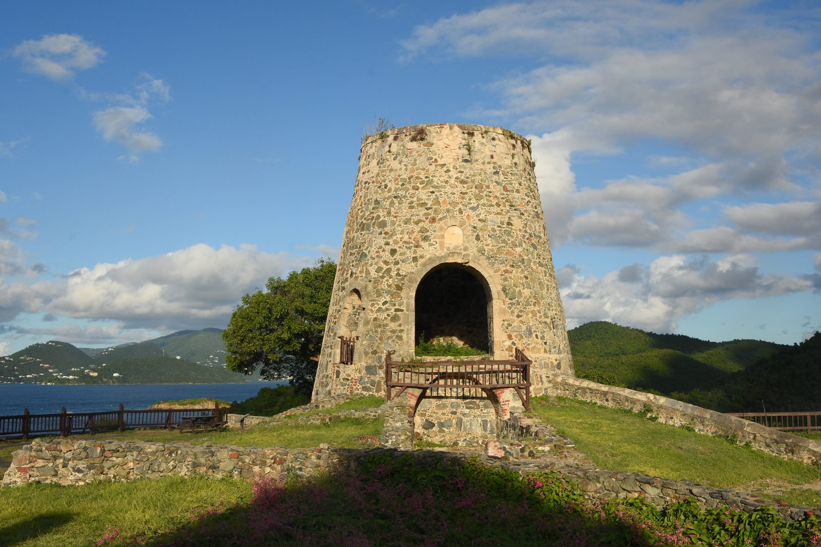 A round stone windmill tower with an arched opening standing on a grassy terrace of low ruined walls, forested green hills and a strip of sea behind it