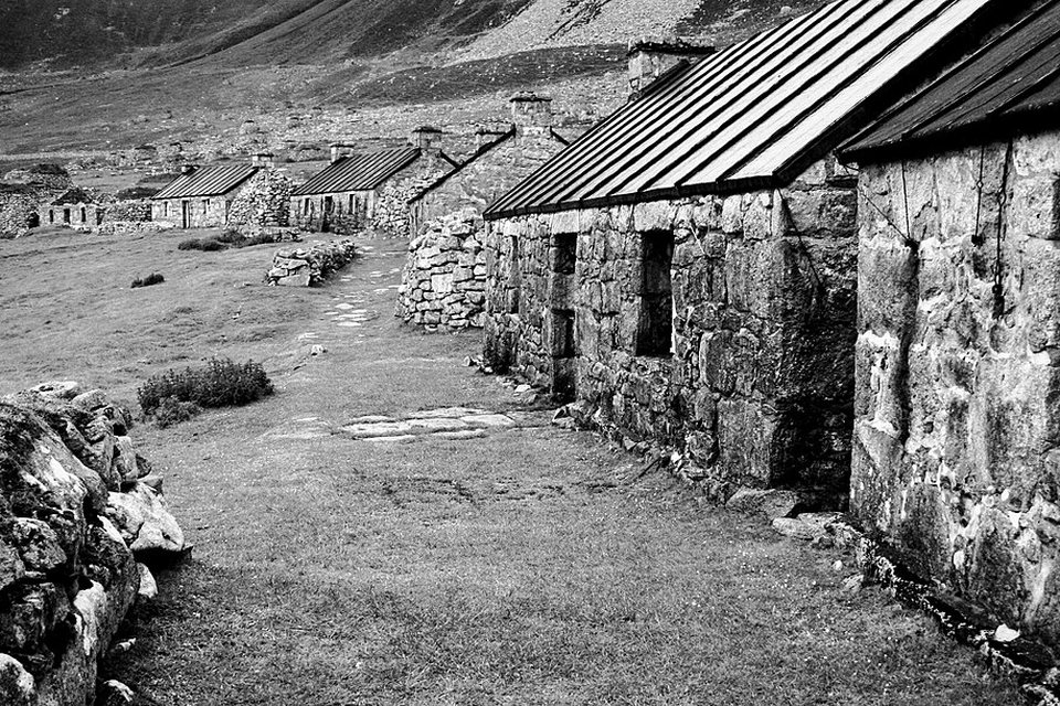A black-and-white view of stone cottages with tin roofs along a path