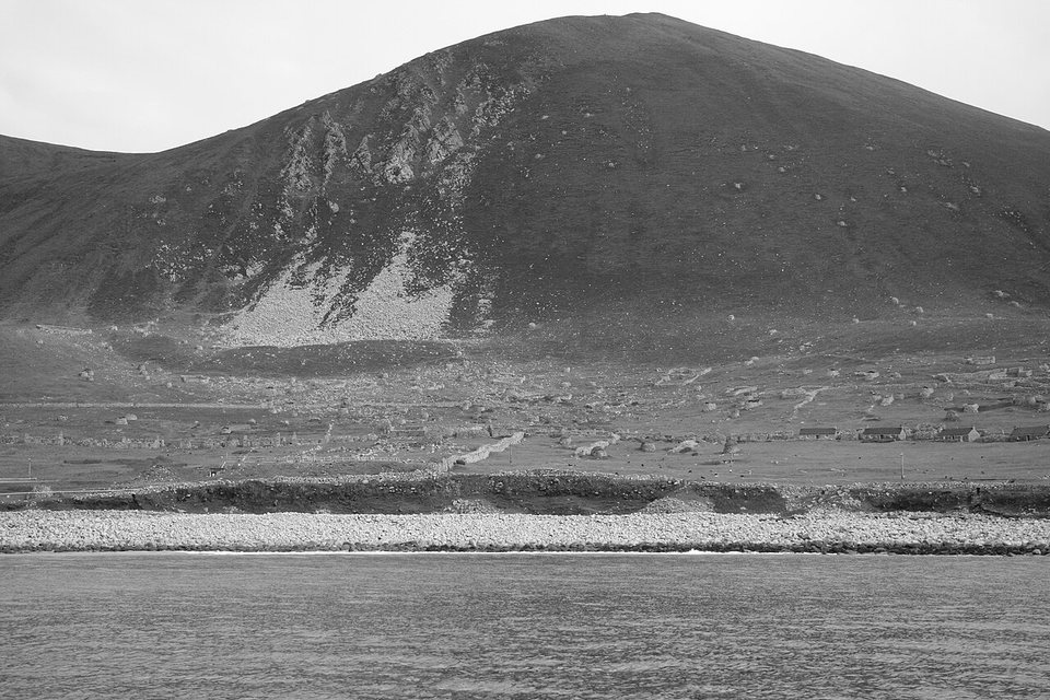 A black-and-white view across water to a slope dotted with storehouses below a high hill