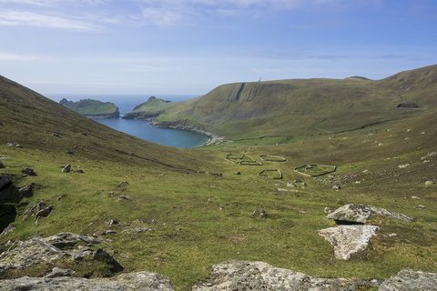 A green bowl of hillside scattered with stone storehouses and walled enclosures above a bay, with a sea stack beyond