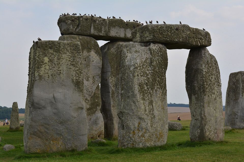 A trilithon of two huge upright stones capped by a lintel with a row of small birds along its top, other stones and a green field behind under a pale grey sky