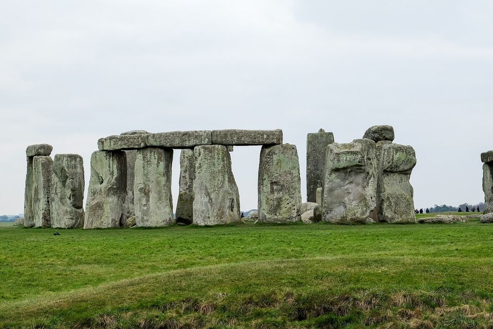 A row of grey standing stones with a long lintel across the top of the centre group on a green field under a flat grey sky