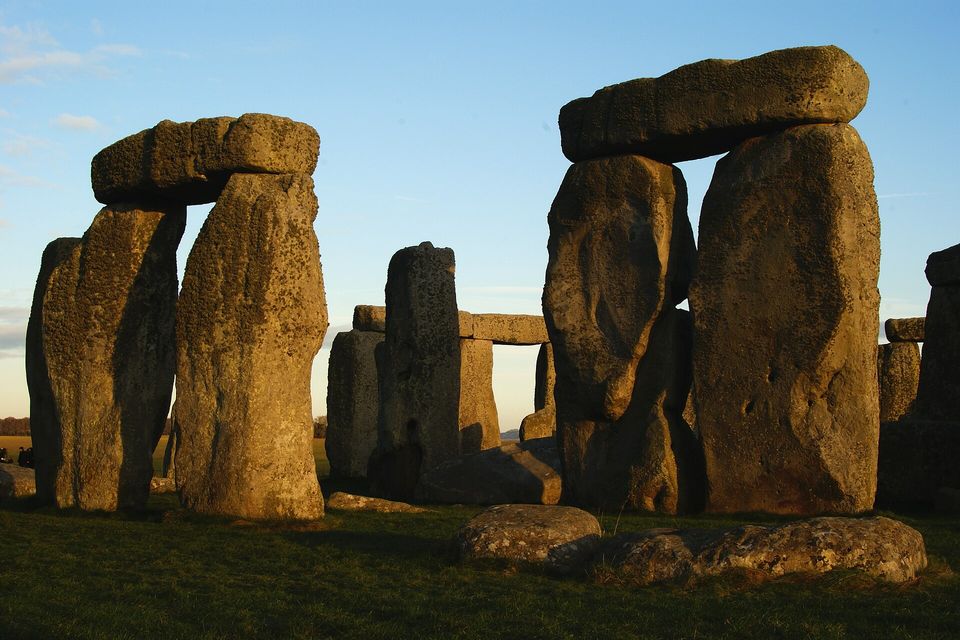 Two trilithons of huge weathered stones with lintels lit gold by low sun against a blue sky, more stones between them and short grass at the bottom