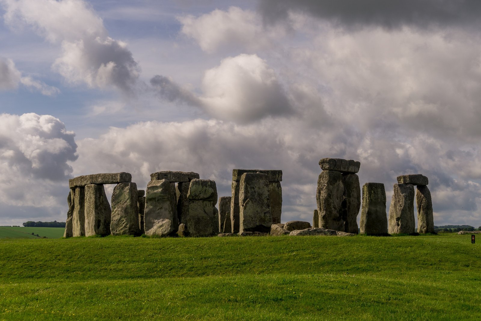 A ring of grey standing stones with lintels on a low green rise under a large sky of white and dark clouds, one small marker post at the right edge