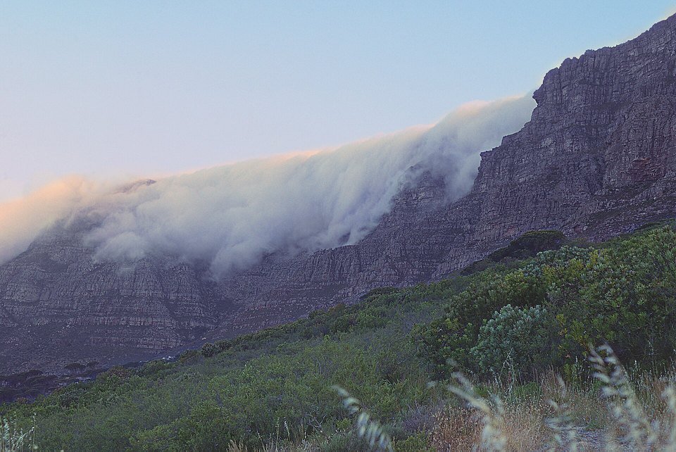 White cloud pouring over the edge of a mountain plateau in low light, with dark cliffs below and green scrub in the foreground