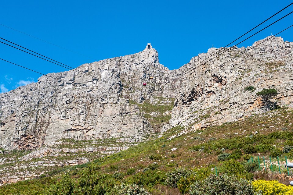 A small red cable car on wires climbing a pale rocky cliff face toward the station at the top, under a deep blue sky