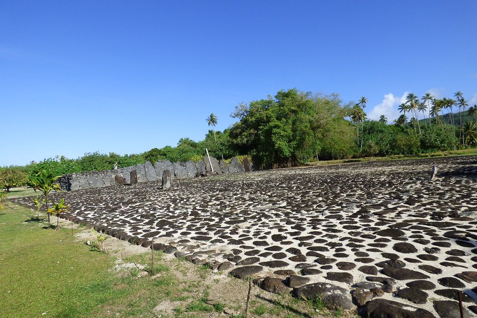 A wide court paved with black stones set in pale coral sand, with a long wall of upright slabs and palm trees at the far side under a deep blue sky