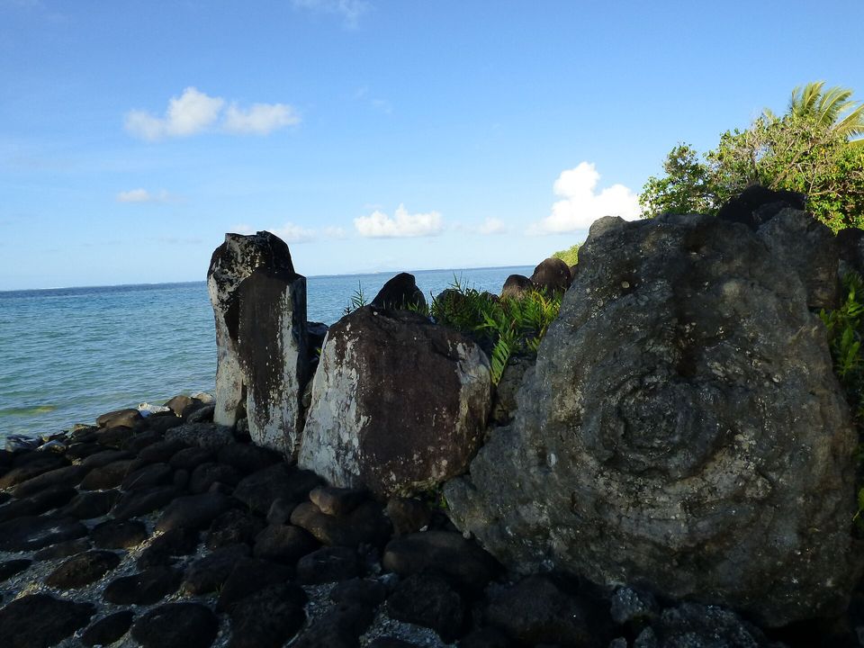 Three tall upright stones, two of them pale, beside a large rounded boulder with a whorled surface, on a black cobbled platform with the sea beyond