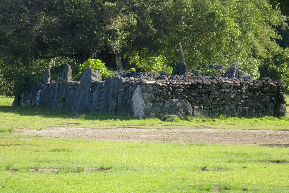 A long low wall of grey stone with upright slabs standing along its top, seen across a lawn with tall trees behind