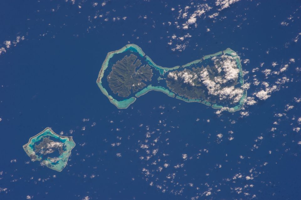 Two green islands joined inside one pale turquoise reef in deep blue ocean, the right one partly under white cloud, and a smaller ringed island at lower left, seen from orbit