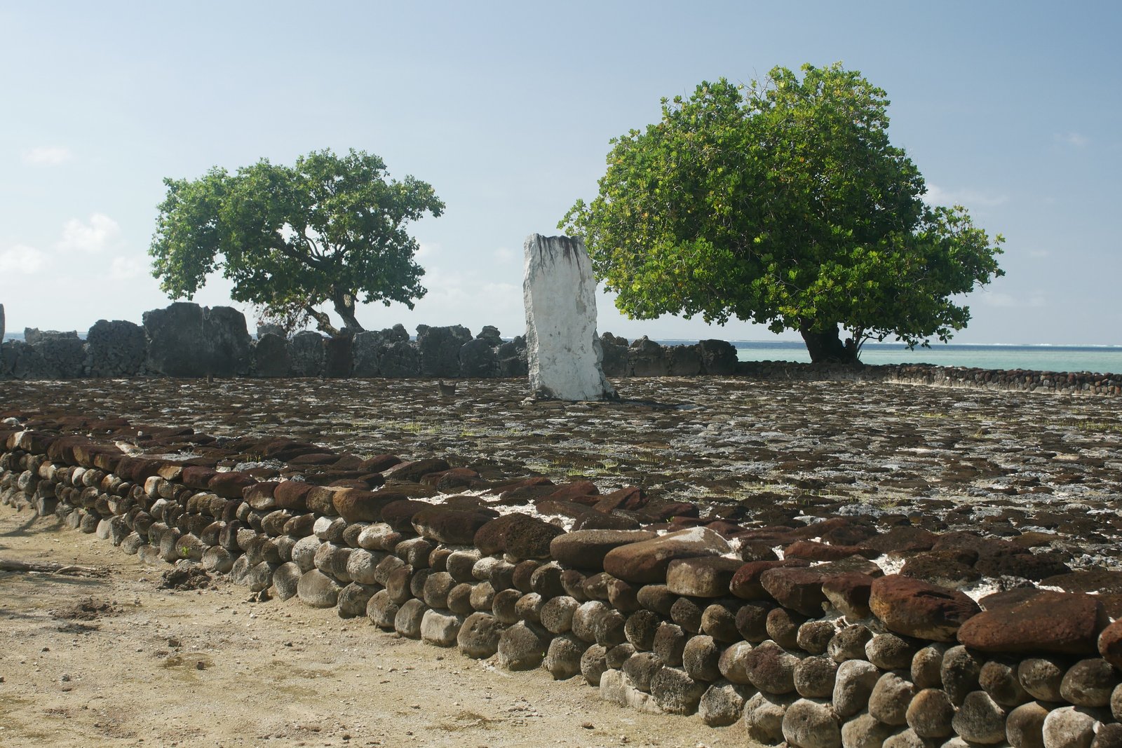 A paved stone court with a single tall white standing stone at its centre, two trees behind it and pale lagoon water beyond a low black wall