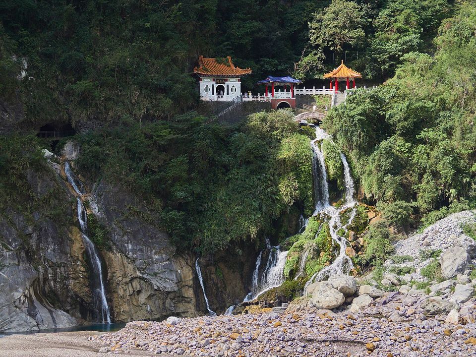 A small white pavilion with orange roofs on a cliff, waterfalls pouring down the rock beneath it