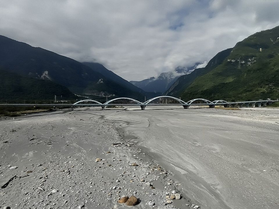 A white arched bridge across a broad grey riverbed, mountains and cloud closing in behind