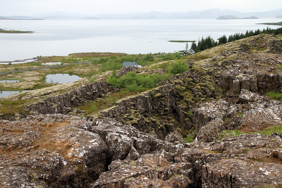 Mossy lava ground split by deep cracks, a few houses and ponds below, a broad pale lake stretching to distant hills