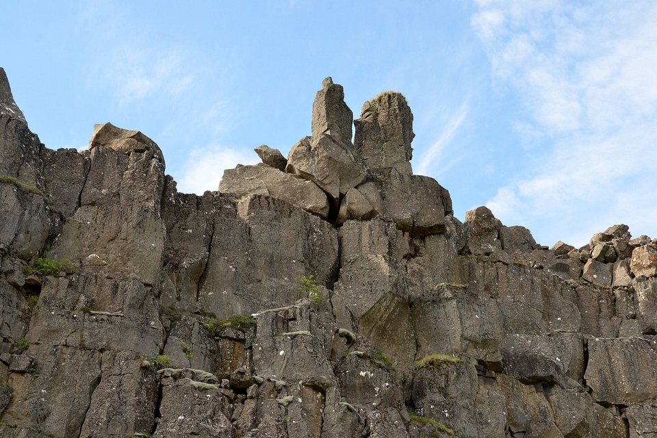 A grey cliff of stacked, columnar lava blocks with a few pinnacles against a blue sky