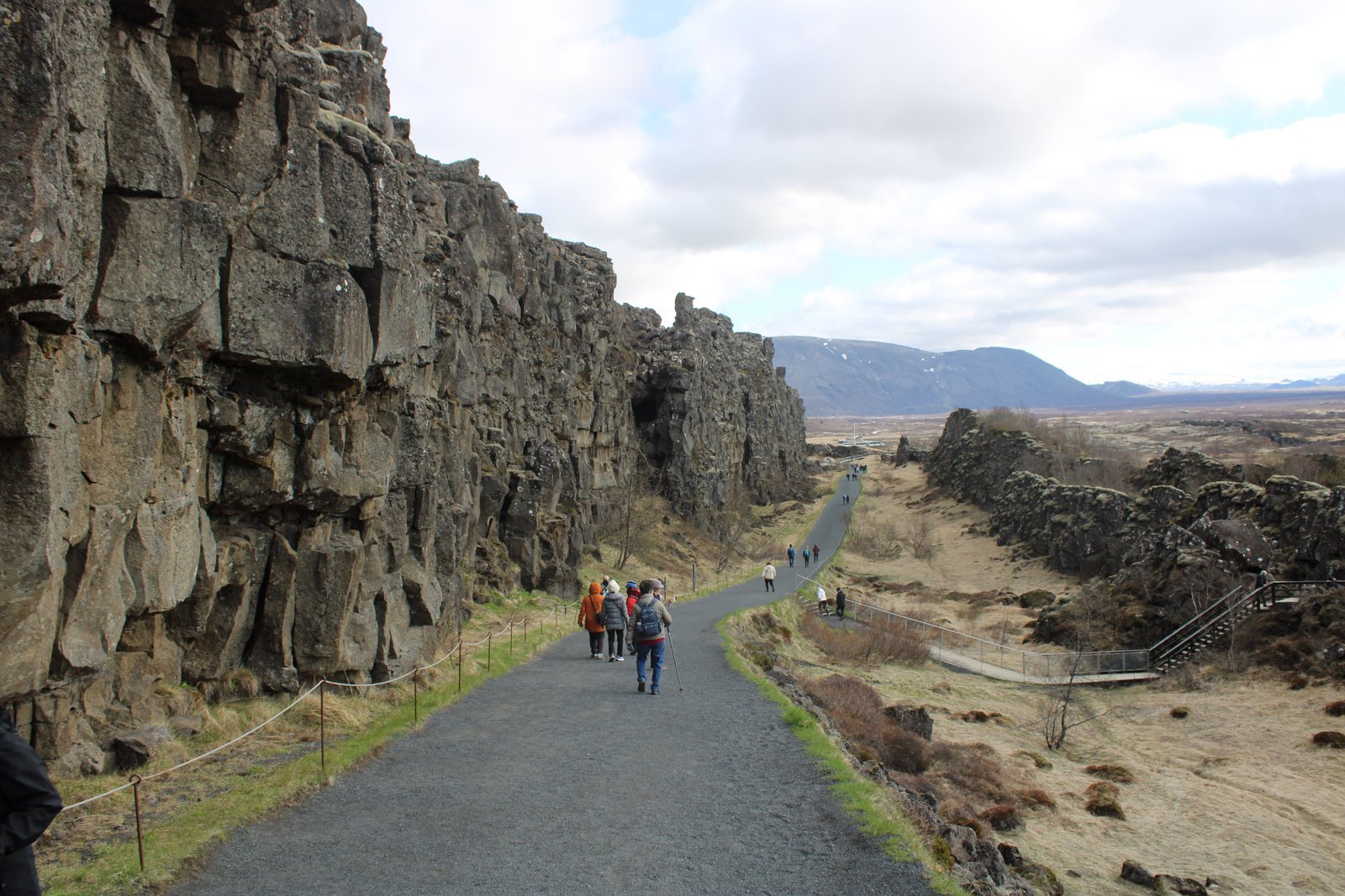 A columnar basalt wall beside a broad gravel path, people walking the valley floor, lowland plain and lake opening out beyond