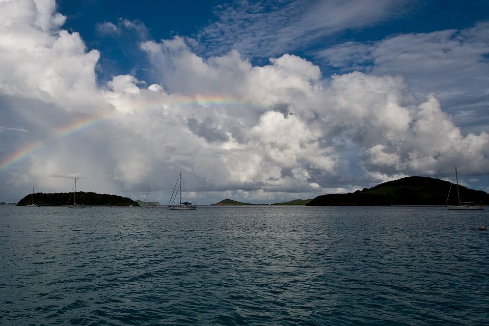 A rainbow arcs through towering white cloud above low green islands and anchored yachts on a dark sea