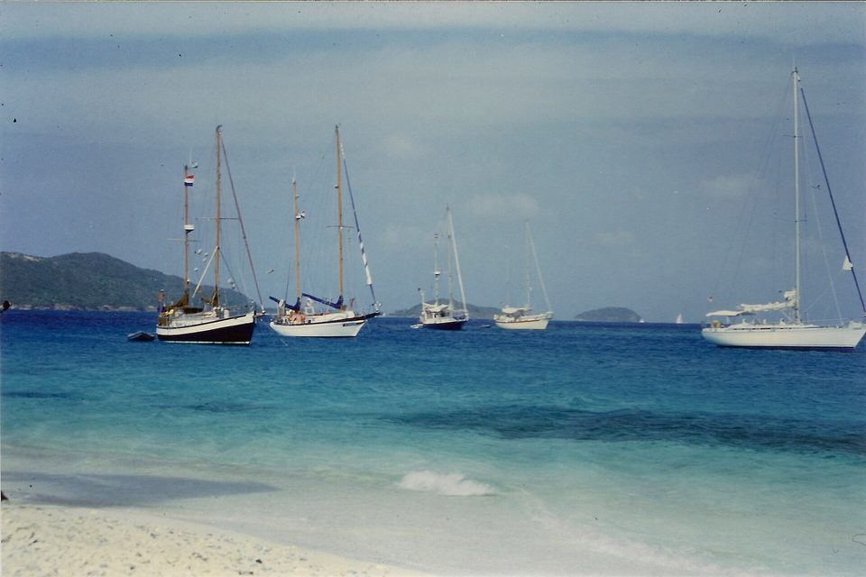 Several wooden and white sailing yachts lie at anchor in turquoise water off a white beach, with green islands behind