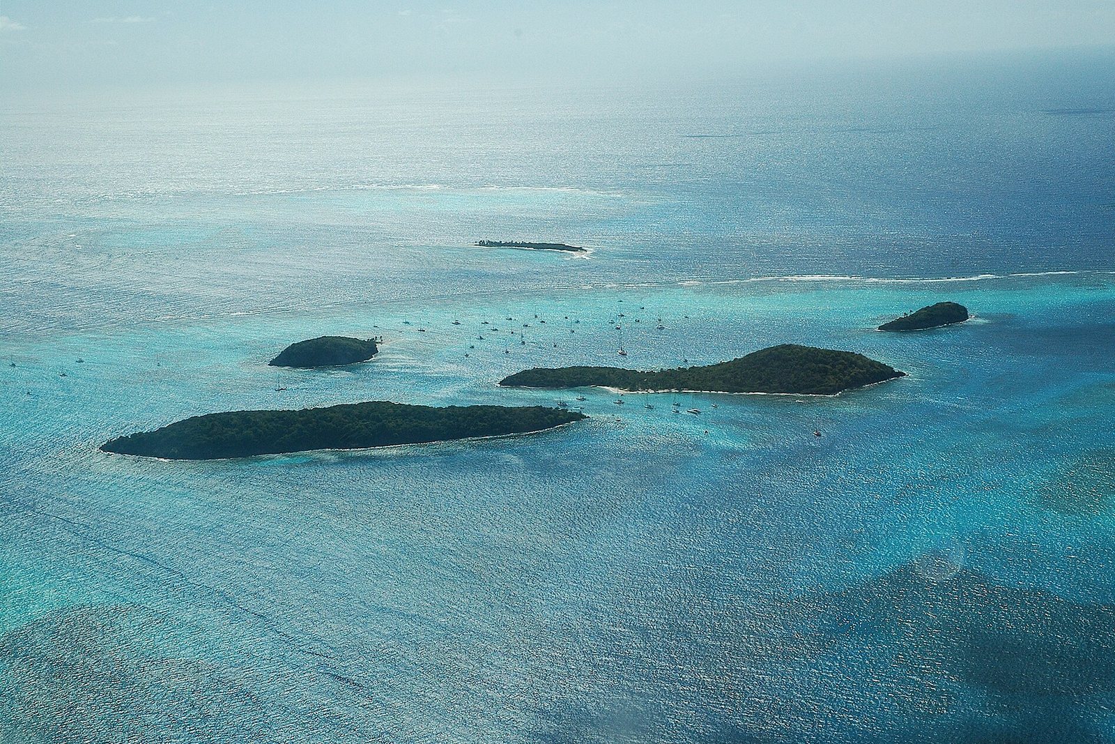Four forested cays sit in pale turquoise shallows with yachts anchored between them and the surf line of the outer reef beyond, under a hazy bright sky