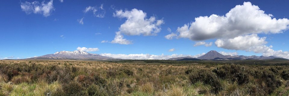 A wide tussock plain under a blue sky with white clouds, a snow-capped broad volcano on the left and a steep symmetrical cone on the right
