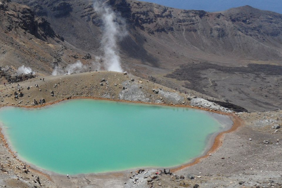 A vivid green crater lake ringed with rust-orange rock in a grey volcanic basin, steam rising from a ridge behind