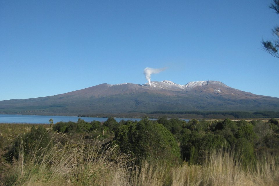 A broad brown volcanic massif with a dusting of snow and a white steam plume, a lake and green scrub in front under a clear blue sky