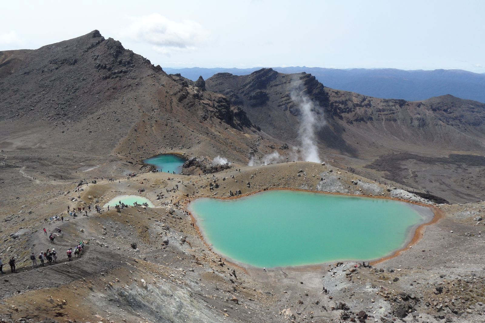 Three crater lakes of vivid green and turquoise ringed with rust-orange rock, steam rising from a ridge behind, and a line of walkers descending grey scree towards them