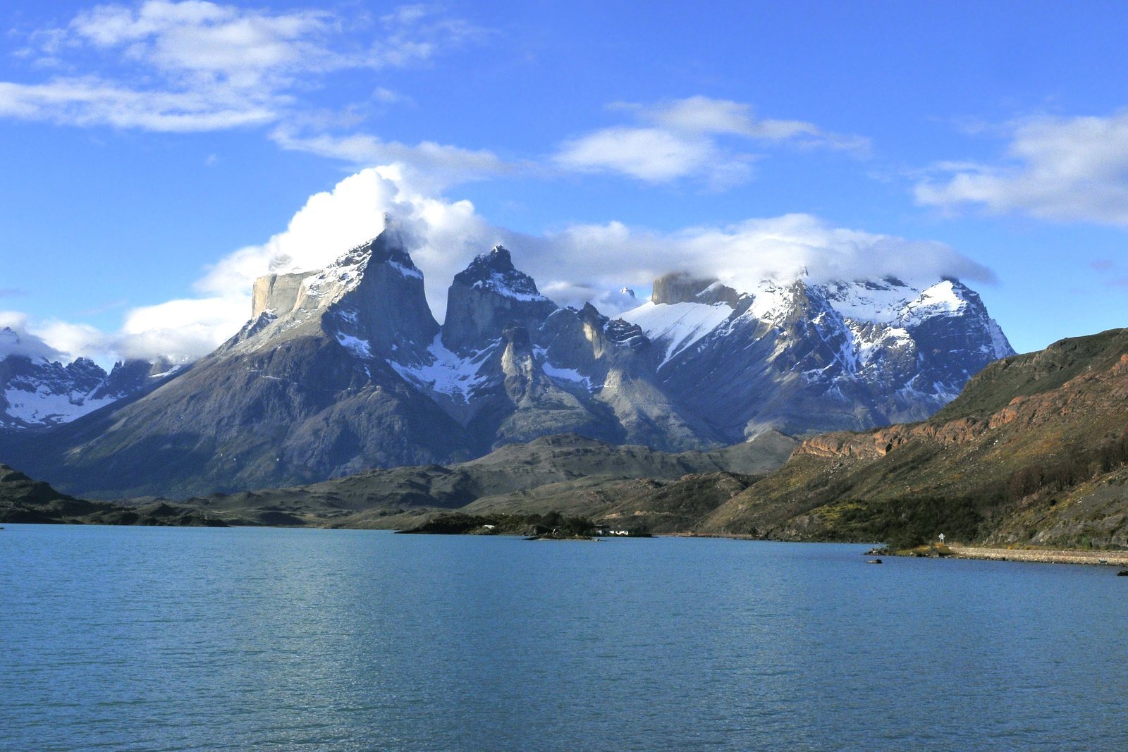 Dark banded rock caps sitting on pale granite peaks with snow in the gullies, cloud spilling over the summits, blue lake in the foreground