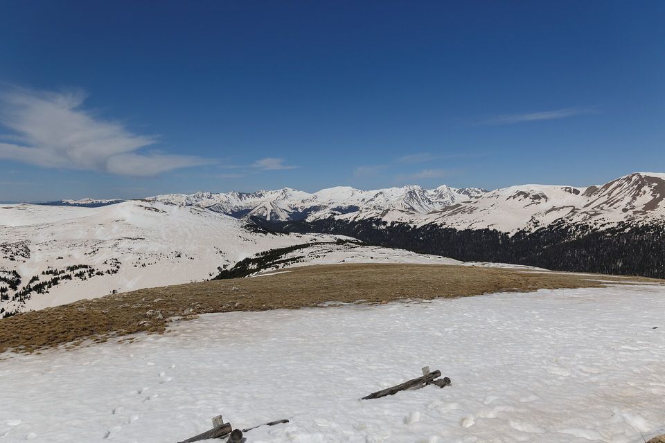 A wide snowfield on open high tundra, a few dead sticks poking through, snow-streaked rounded mountains along the far horizon under a deep blue sky
