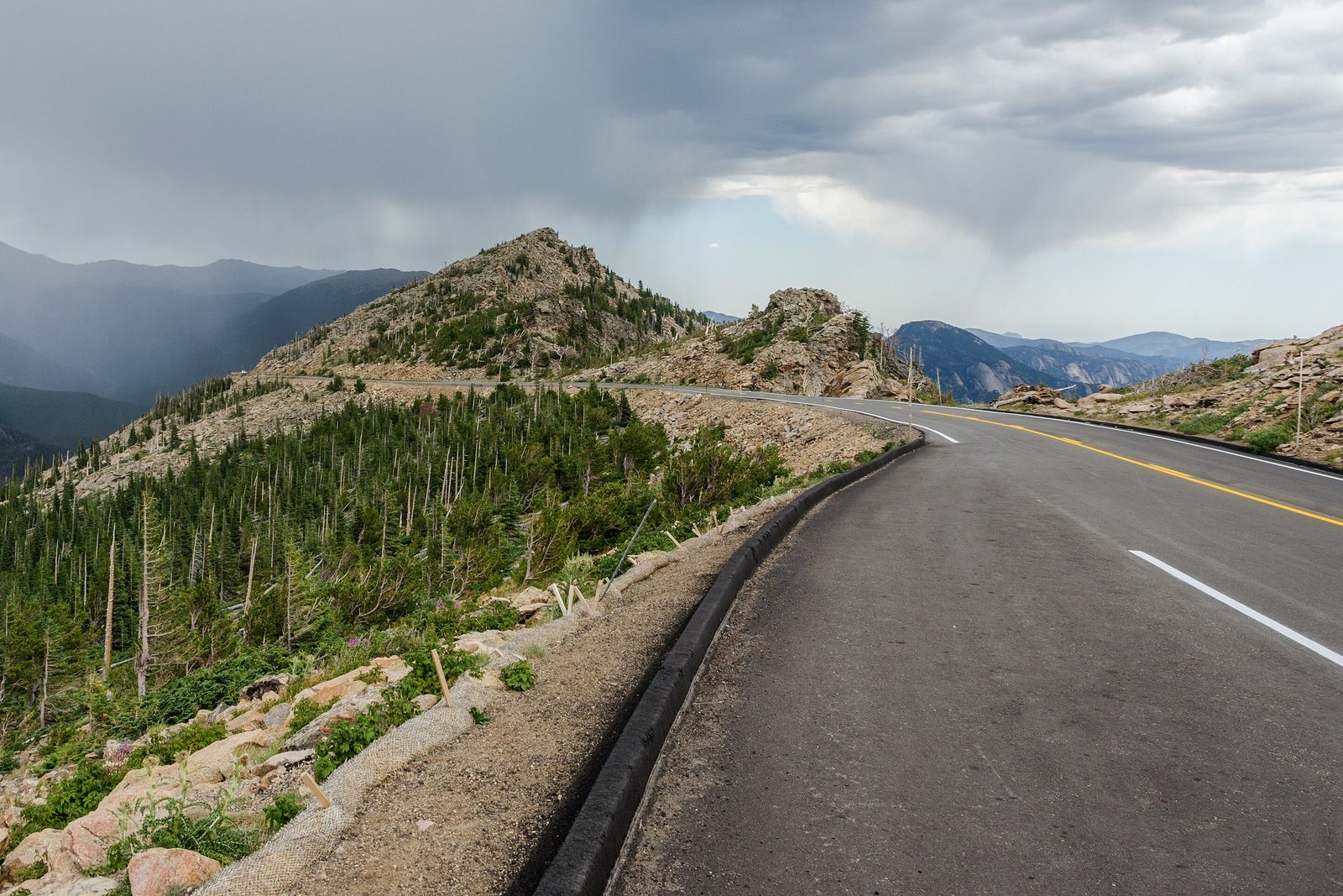 Trail Ridge Road curving along a rocky ridge above treeline toward a pointed knob, rain falling from dark cloud over the mountains at the left