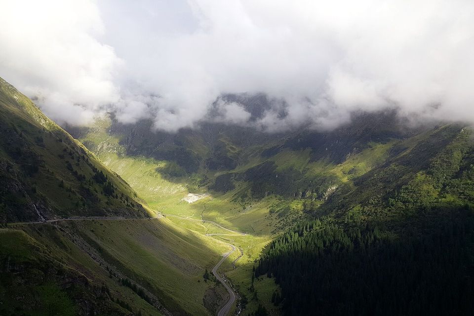 Low cloud spilling over a green glacial valley, a thin road winding along the valley floor and sunlight catching the slopes