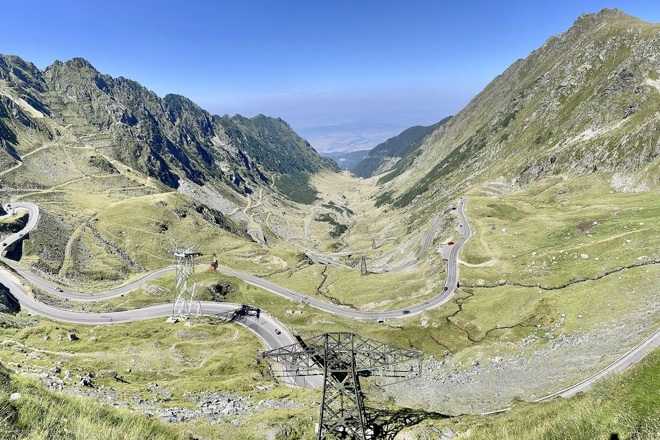 The whole northern descent laid out below the pass: loops of road across a green valley between rocky ridges, pylons of a cable car in the foreground