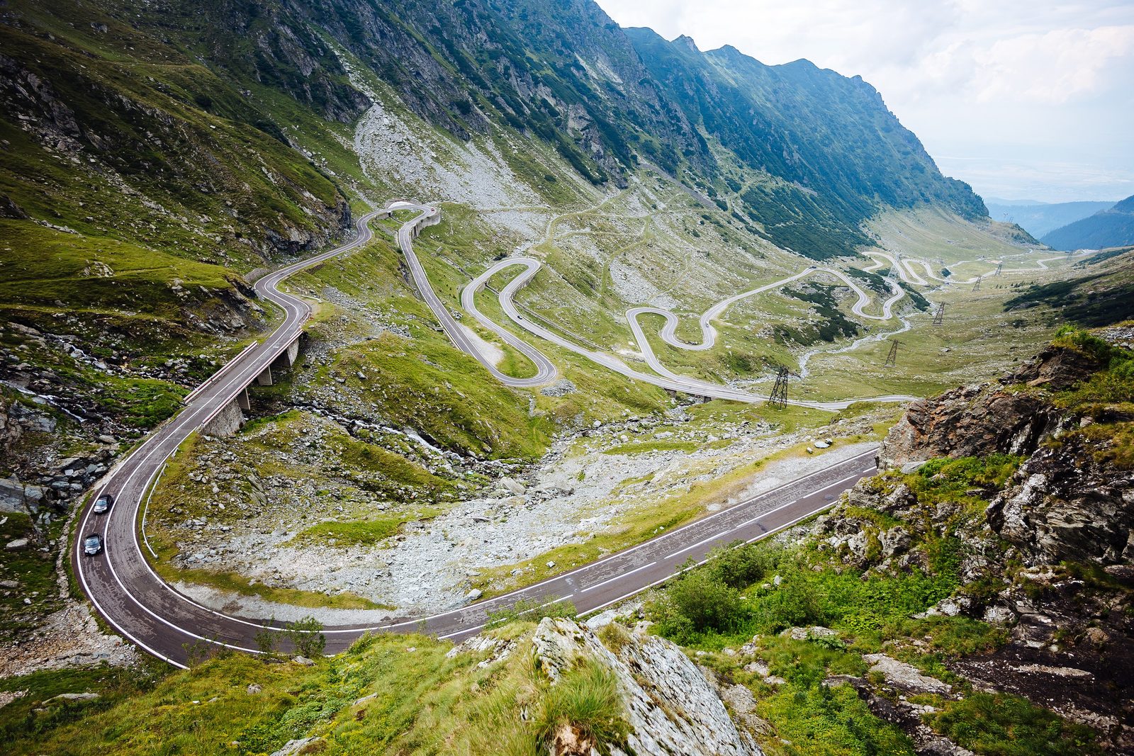 The Transfăgărășan’s hairpins stacked up a green glacial valley beneath grey Carpathian ridges, a viaduct in the foreground and pylons climbing the slope