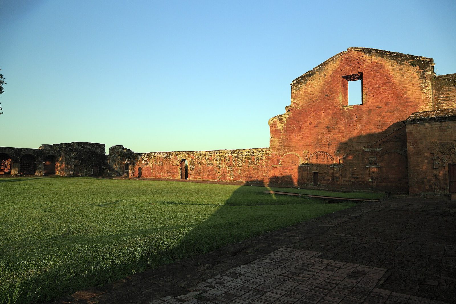 The west wall and gable of a ruined church glowing red in low sun across a cloister lawn, carved arches under the cornice