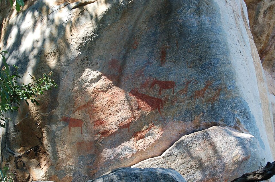 Red animal figures across a large boulder face in dappled shade