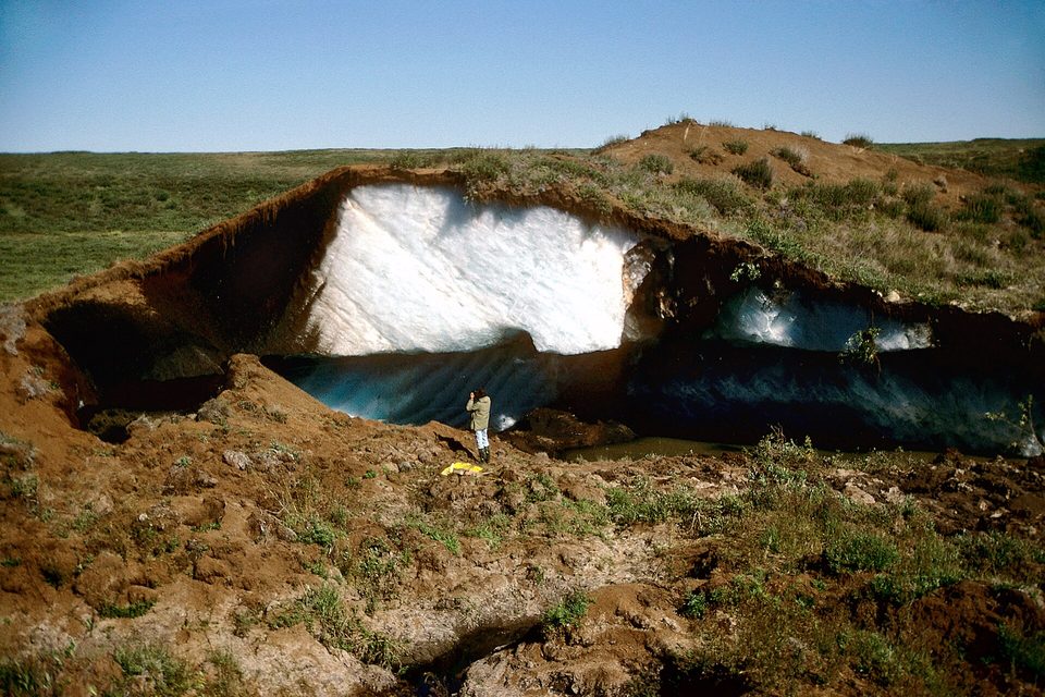 The cut face of a pingo showing a wall of solid ice under a layer of dark peat, a small figure standing at its foot for scale