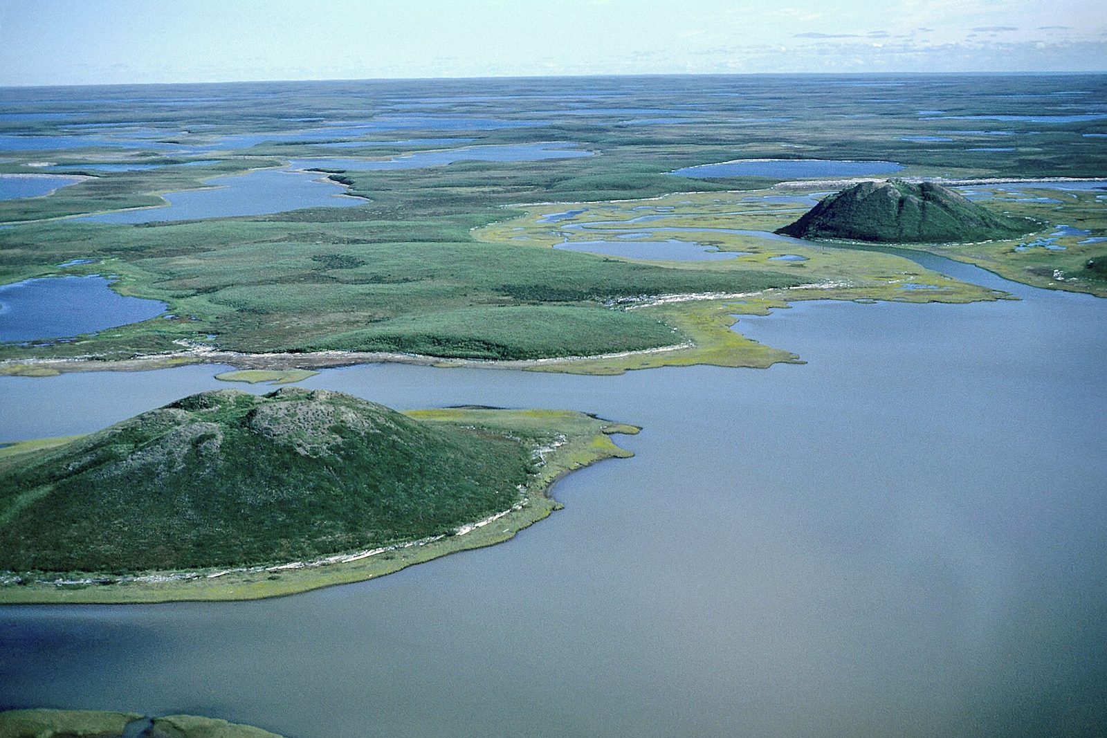 Two green ice-cored hills rising from drained lakes on flat tundra, with water running to the horizon