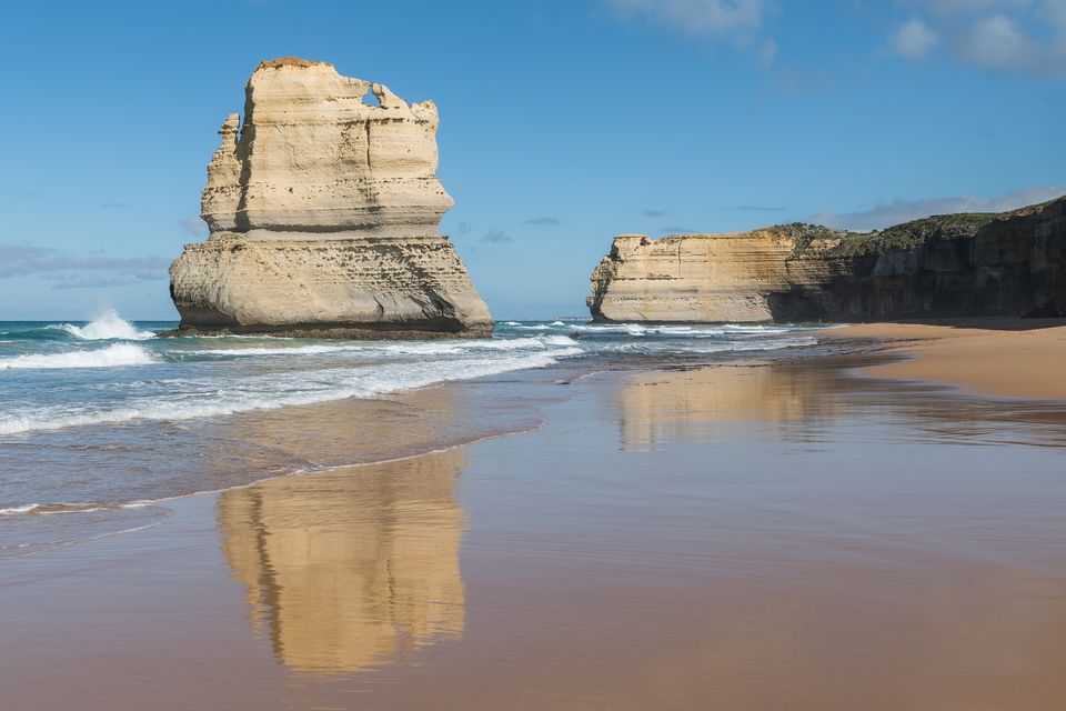A wide, pale limestone stack rising from a shining wet beach and reflected in it, with a cliff along the shore behind under a blue sky