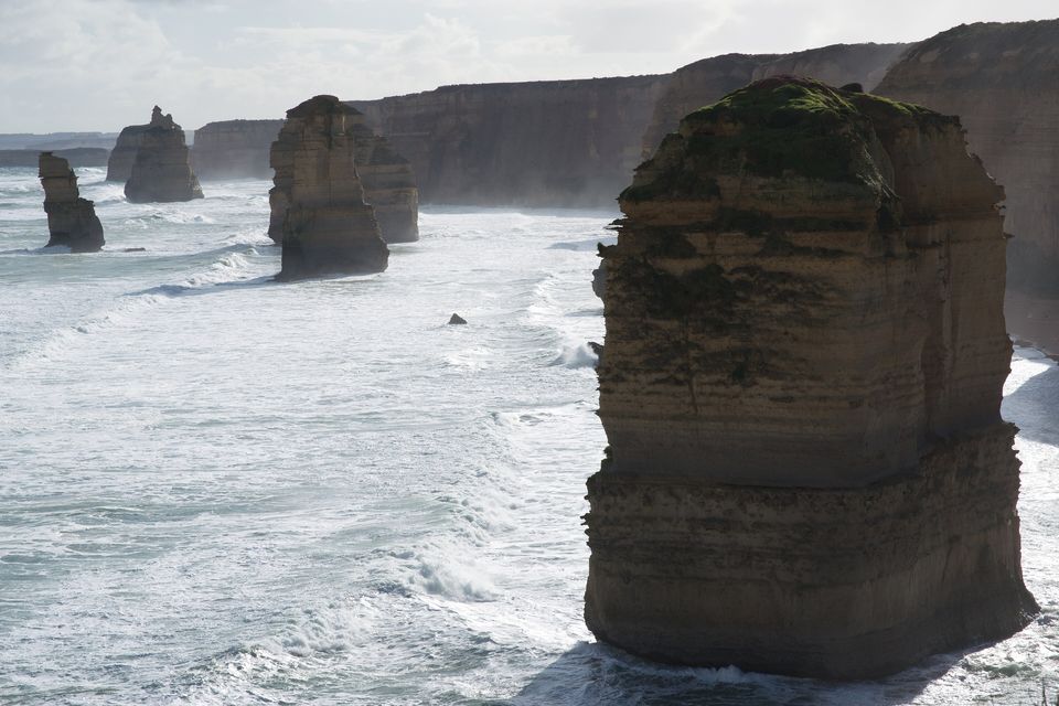 A square, dark-topped stack in the foreground banded with horizontal layers of limestone, more stacks and a line of cliffs fading into sea spray beyond