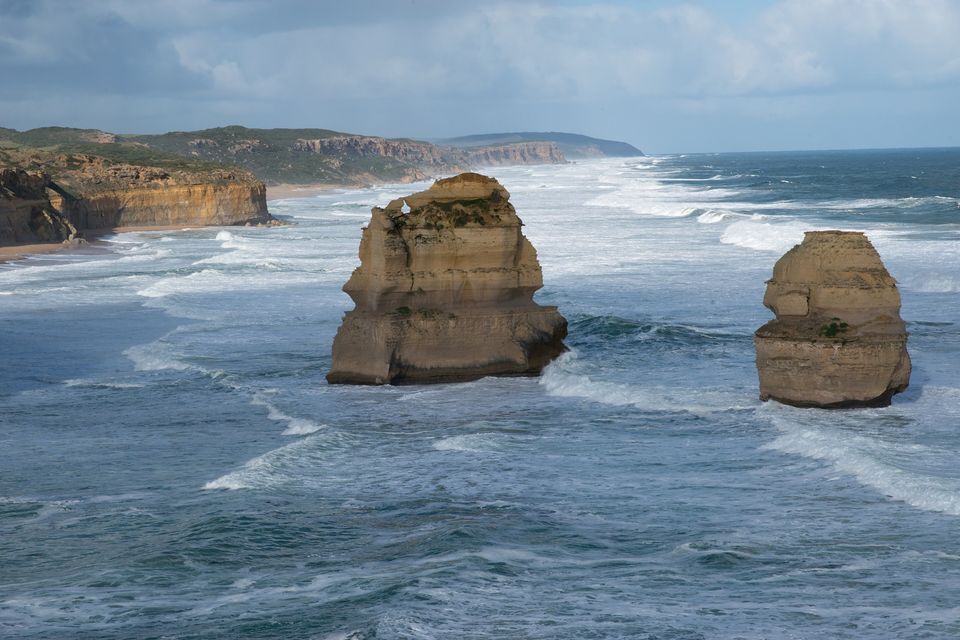 Two pale limestone stacks standing in rough grey-blue surf off a long line of cliffs and low headlands