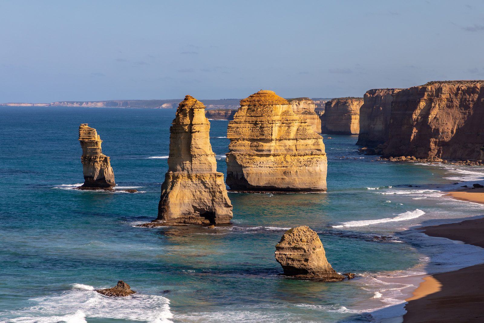 Golden limestone stacks standing in a deep blue sea below dark-topped cliffs, with surf and a sandy beach at the right under a pale blue sky