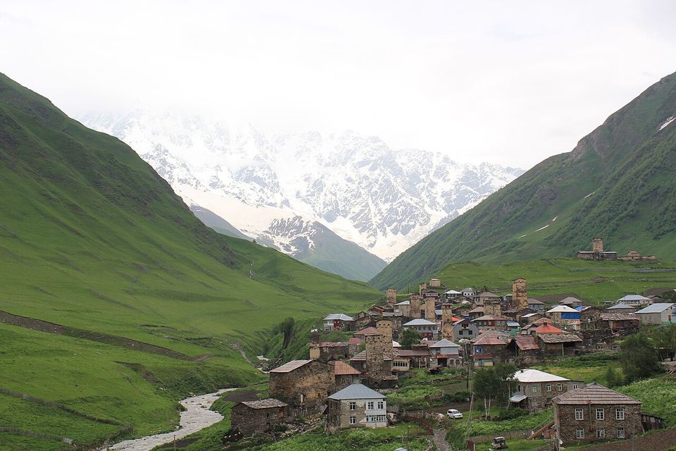 A village of stone towers and houses on green slopes beside a river, snow peaks rising behind