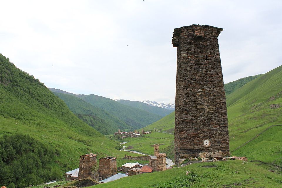 A tall dark stone tower with no ground-floor opening on a grassy slope, a village of towers below