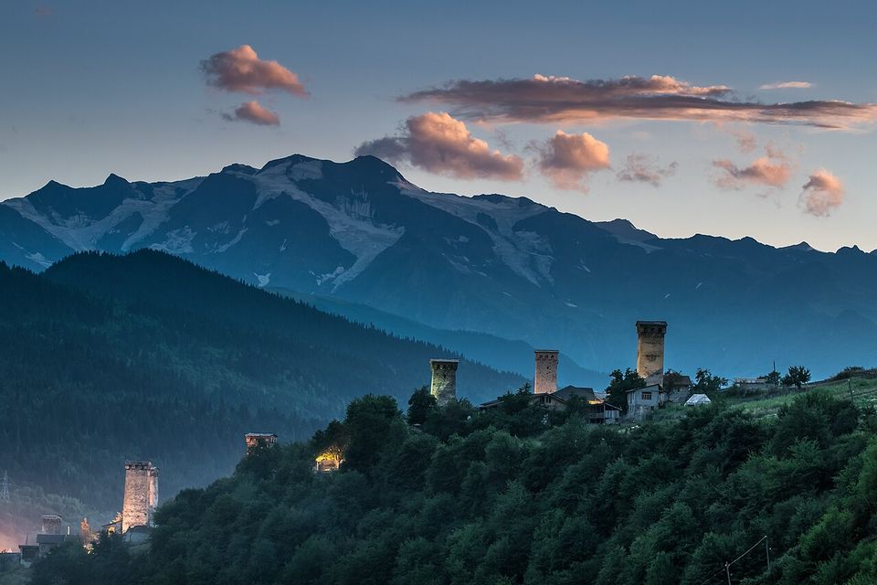 Several stone towers on a wooded ridge at dusk, a snow-streaked mountain wall and pink clouds behind