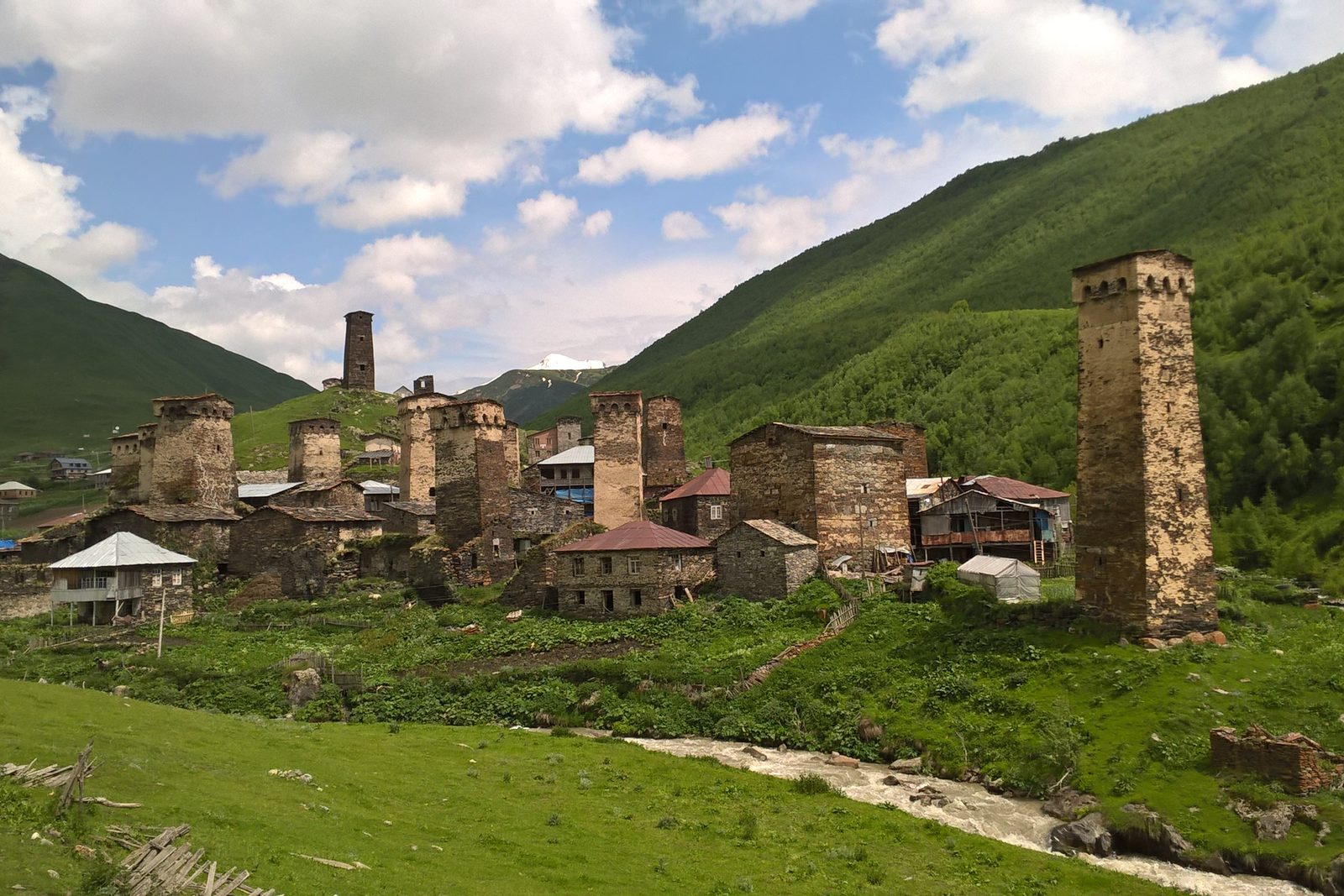 A cluster of stone defensive towers rising among slate-roofed houses on a green valley floor, steep grassy slopes closing in on both sides and a snow patch in the gap