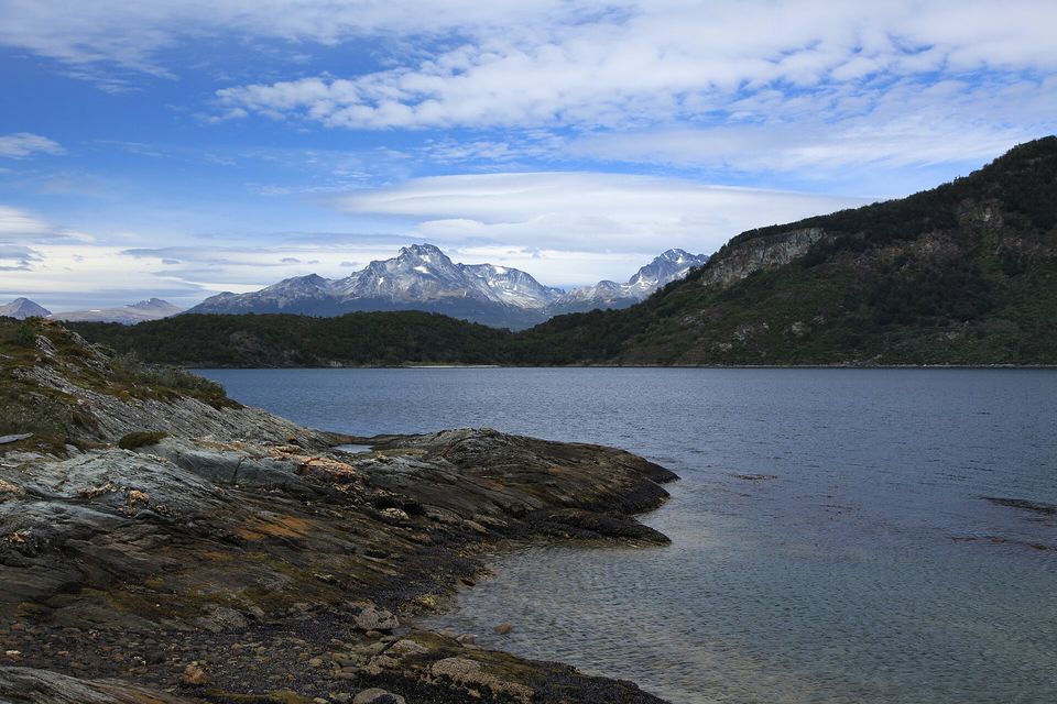 A dark bay with a rocky lichen-covered shore in the foreground, forested hills and a snow-dusted mountain beyond under scattered cloud