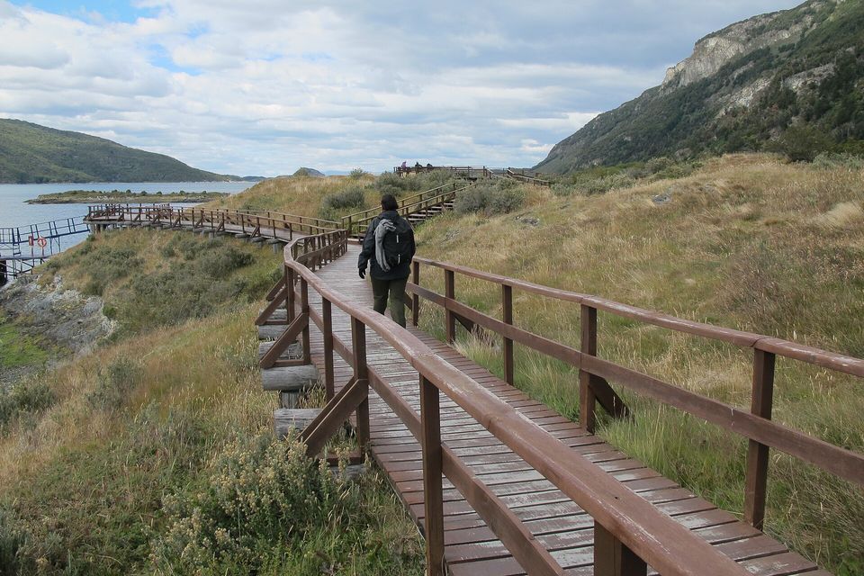 A wooden boardwalk curving along a grassy shore beside a bay, one walker seen from behind, forested hills at both sides